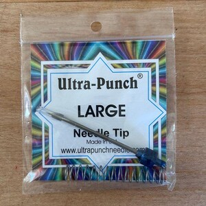 May include: A clear plastic package containing an Ultra-Punch needle tip. The package has a colorful geometric design and the words "Ultra-Punch" and "LARGE" are visible. The needle tip is silver with a blue handle and a metal spring.
