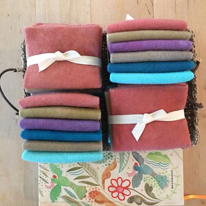 May include: Stacks of colorful felt squares in shades of pink, purple, blue, and tan, tied with white ribbon. The felt is arranged in a woven basket, with a decorative book featuring a floral and bird design visible below.