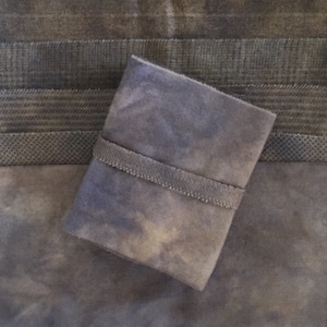 May include: A small, square, gray fabric pouch with a stitched band detail. The pouch is set against a backdrop of various gray fabrics with different textures and patterns, creating a layered effect.