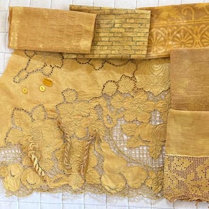 May include: An assortment of vintage textiles in shades of yellow and gold. The collection includes a floral lace tablecloth, a brick-patterned fabric, and other textured materials. Several small buttons and beads are also visible.