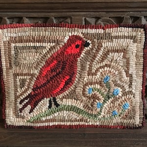 May include: Handmade rug featuring a red bird perched on a green branch with blue flowers. The bird has a black eye and is set against a neutral background. The rug is framed with a red border and has a textured, handcrafted appearance.