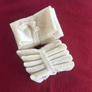 May include: Two stacks of off-white, rectangular cloths, each bound with a matching fabric strip. One stack is neatly folded, while the other is a slightly fanned arrangement. The cloths rest on a deep red, textured surface.