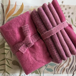 May include: A collection of pink wool fabric bundles, some rolled and tied with a ribbon, and others folded and secured with a bow. The fabrics vary in shades of pink, from light to deep rose, and are arranged on a patterned background.