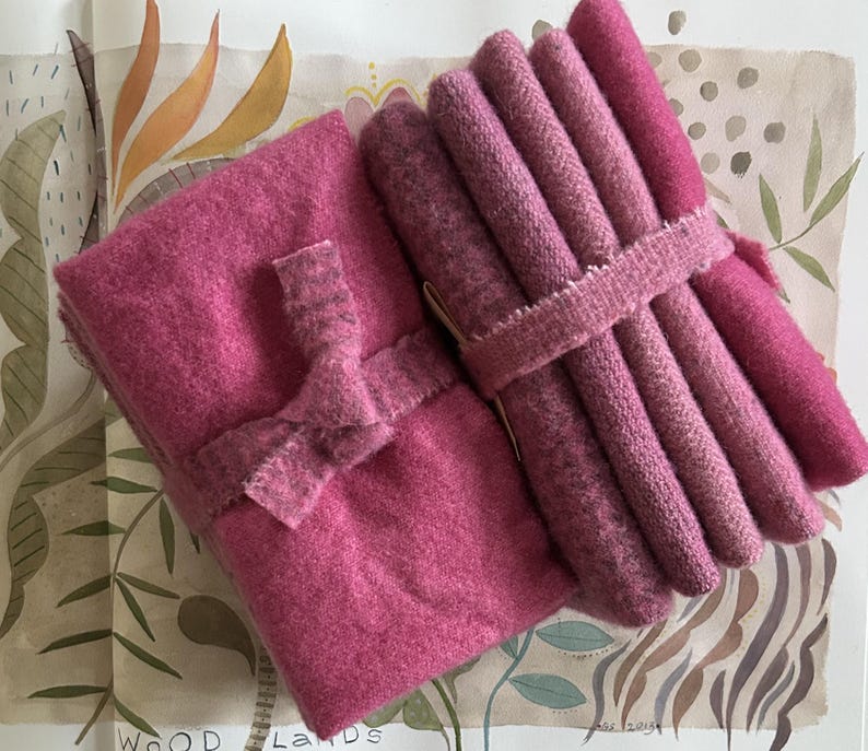 May include: A collection of pink wool fabric bundles, some rolled and tied with a ribbon, and others folded and secured with a bow. The fabrics vary in shades of pink, from light to deep rose, and are arranged on a patterned background.
