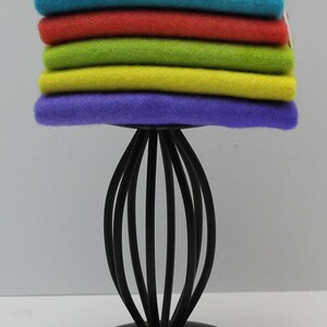 May include: A stack of folded, colorful sweaters in shades of pink, teal, red, green, yellow, and purple. The sweaters are displayed on a black metal stand with a decorative, curved design. The background is plain white.
