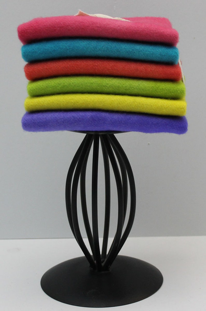 May include: A stack of folded, colorful sweaters in shades of pink, teal, red, green, yellow, and purple. The sweaters are displayed on a black metal stand with a decorative, curved design. The background is plain white.