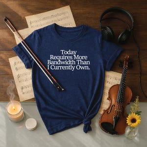 Puede incluir: Camiseta azul marino con el texto "Today Requires More Bandwidth Than I Currently Own." Un violín, arco, partituras, auriculares, una vela y un pequeño jarrón con un girasol están dispuestos sobre una superficie de madera.