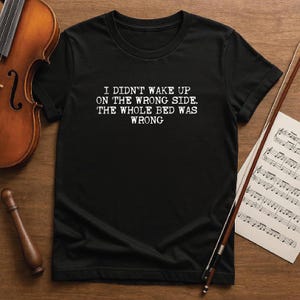 Puede incluir: Camiseta negra con el texto blanco: "I DIDN'T WAKE UP ON THE WRONG SIDE. THE WHOLE BED WAS WRONG." Un violín, un arco y partituras son visibles sobre una superficie de madera.