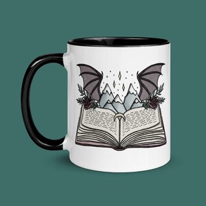 May include: White ceramic mug with a black rim and handle. The mug features a black and white illustration of a book with mountains and a pair of wings on top.