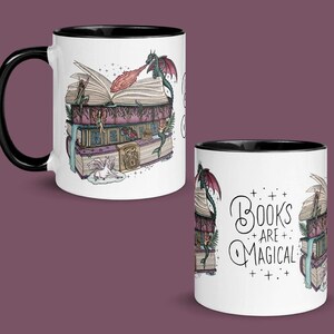 Könnte beinhalten: Drei weiße Keramikbecher mit schwarzen Rändern. Die Becher zeigen ein Design mit einem Drachen, der über einem Stapel Bücher fliegt. Der Text "Books are magical" steht auf einem der Becher.