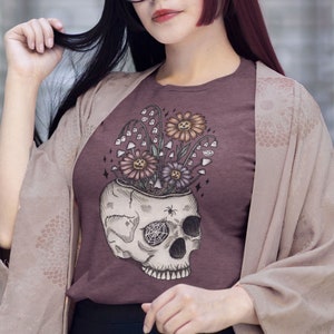May include: A woman wearing a maroon t-shirt with a skull graphic featuring flowers growing out of the top of the skull. The skull has a spiderweb over one eye socket. The woman is wearing a long, light brown cardigan and a black skirt.