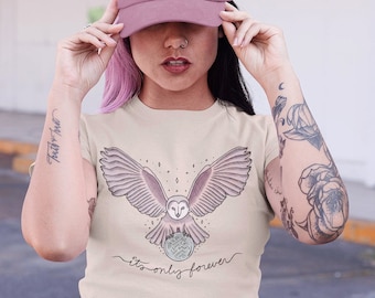 May include: A woman wearing a beige t-shirt with a graphic of an owl flying over a moon with the text "it's only forever". She is also wearing a pink baseball cap and blue jeans.