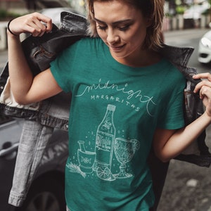 May include: Teal t-shirt with the words "midnight margaritas" in a script font. Below the text is a white illustration of a bottle, glass, mortar and pestle, and lime slices. The shirt is being worn.
