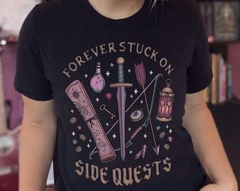 May include: Black t-shirt with the text "FOREVER STUCK ON SIDE QUESTS" and a graphic of a sword, bow and arrow, potion bottle, scroll, key, and lantern. The design is in shades of pink, purple, and teal.