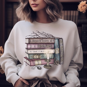 Puede incluir: Una sudadera blanca con un gráfico de una pila de libros, una vela encendida y un reloj de bolsillo. Los libros incluyen títulos como "Cumbres Borrascosas", "Jane Eyre" y "La inquilina de Wildfell Hall".