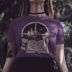 May include: A woman wearing a purple t-shirt with a graphic of an open book with a window and moon in the background. The book has text on the pages and the window has a crescent moon and stars. The woman is standing in a forest.