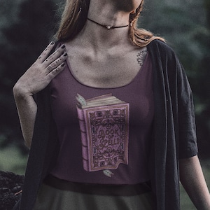 Puede incluir: Una mujer con una camiseta de tirantes de color morado oscuro con un gráfico de un libro de hechizos, una falda verde oscuro y una chaqueta negra. Está de pie en un entorno de bosque.