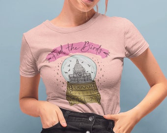 Puede incluir: Camiseta rosa claro con el texto "Feed the Birds" sobre un diseño de bola de nieve con un edificio y pájaros. La modelo lleva una falda vaquera negra.