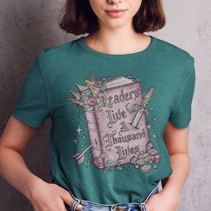 May include: A teal green t-shirt with a graphic of a book with the text "Readers Live a Thousand Lives". The book is surrounded by flowers, a crown, a sword, and other decorative elements.