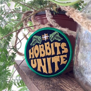 Könnte beinhalten: Ein runder bestickter Aufnäher mit schwarzem Hintergrund und grünem Rand. Der Text HOBBITS UNITE ist mit goldenem Garn gestickt. Über dem Text befindet sich ein weißes vierzackiges Sternsymbol, flankiert von zwei grünen Blattillustrationen.