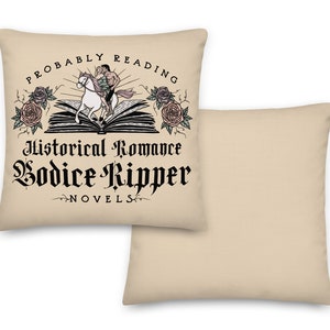 May include: Beige throw pillow featuring a black and white illustration of a person riding a horse on top of an open book. The text "Probably Reading Historical Romance Bodice Ripper Novels" is printed on the pillow.