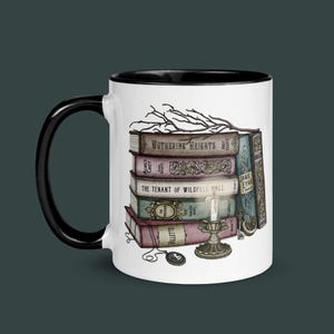 Puede incluir: Taza de cerámica blanca con borde y asa negros. La taza presenta una pila de libros con títulos como "Wuthering Heights", "Agnes Grey", "The Tenant of Wildfell Hall" y "Jane Eyre". También se representan una vela y una llave en la taza.