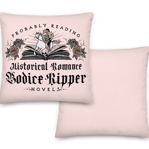 May include: Two pink throw pillows. One pillow features a black and white graphic of a couple riding a horse on top of an open book. The graphic is surrounded by roses and the text "Probably Reading Historical Romance Bodice Ripper Novels".