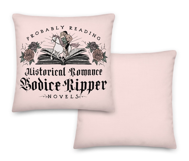 May include: Two pink throw pillows. One pillow features a black and white graphic of a couple riding a horse on top of an open book. The graphic is surrounded by roses and the text "Probably Reading Historical Romance Bodice Ripper Novels".