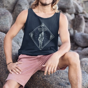 May include: A man wearing a dark blue tank top with a white graphic of a twisted rock and the text "Wandering Wizards Society". He is wearing pink shorts and sitting on a large rock.