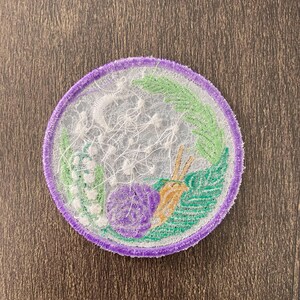 May include: A purple and green embroidered patch with a white floral design and a snail. The patch is circular and has a white background.