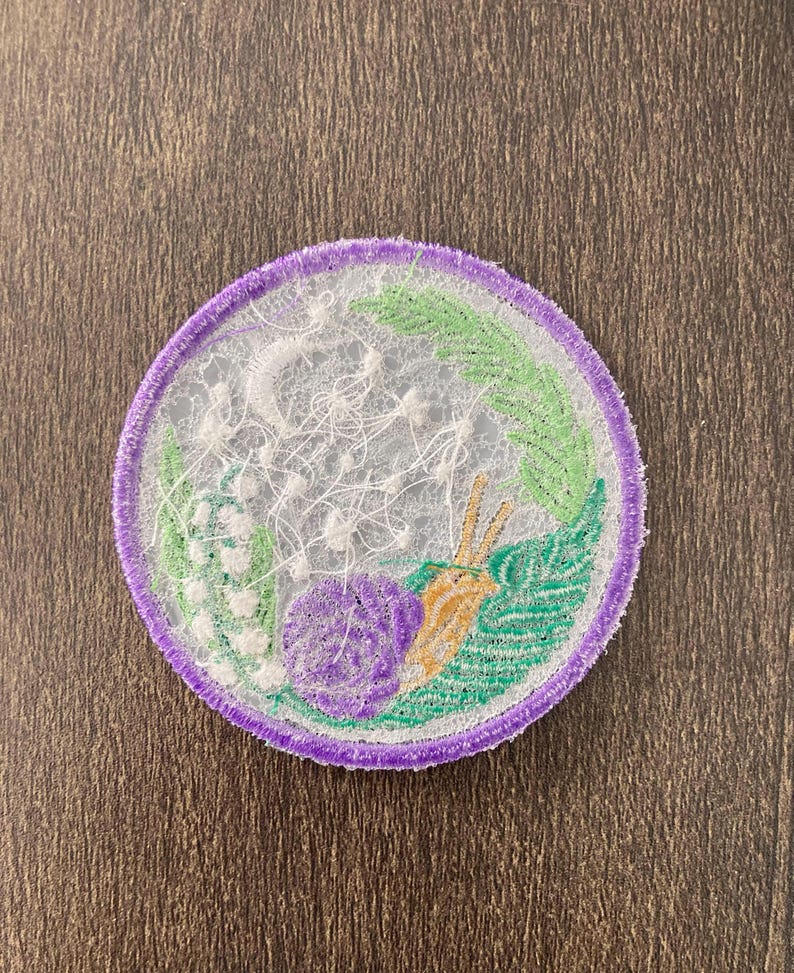 May include: A purple and green embroidered patch with a white floral design and a snail. The patch is circular and has a white background.