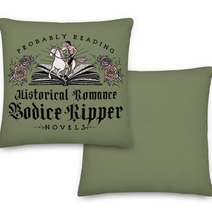 May include: Two green throw pillows, one with a graphic of a couple riding a horse on a book with the text "Probably Reading Historical Romance Bodice Ripper Novels" and the other plain.