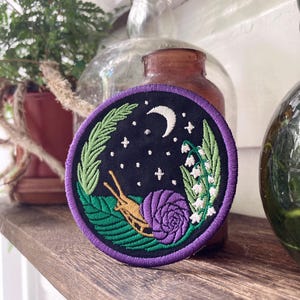 May include: A purple embroidered patch with a snail, a rose, lily of the valley flowers, and a crescent moon on a black background.