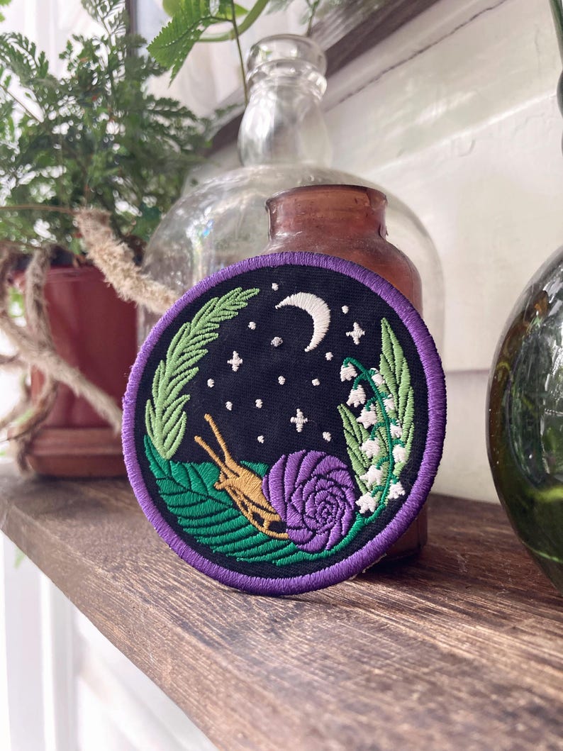 May include: A purple embroidered patch with a snail, a rose, lily of the valley flowers, and a crescent moon on a black background.