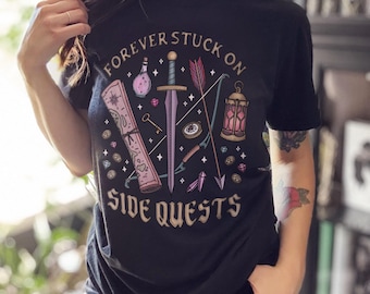 May include: Black t-shirt with the text "FOREVER STUCK ON SIDE QUESTS" and fantasy-themed illustrations. The design includes a sword, bow and arrow, potion bottle, map, key, and gems. The shirt is a crew neck.