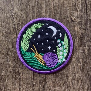 May include: A purple and gold embroidered patch with a snail, leaves, flowers, and a crescent moon on a black background. The patch is surrounded by a purple border.