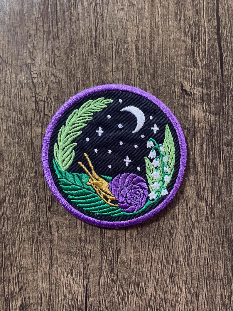 May include: A purple and gold embroidered patch with a snail, leaves, flowers, and a crescent moon on a black background. The patch is surrounded by a purple border.
