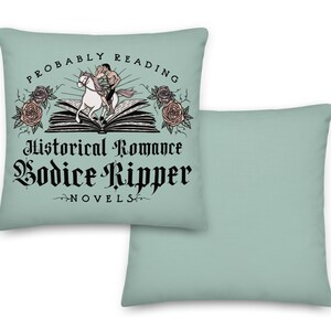 May include: Two decorative throw pillows with a light blue background. One pillow features a black and white illustration of a couple riding a horse on top of an open book with the text "Probably Reading Historical Romance Bodice Ripper Novels".