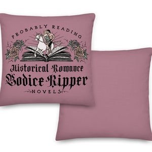 May include: A pink throw pillow featuring the text "Probably Reading Historical Romance Bodice Ripper Novels" in black lettering. The text is surrounded by a graphic of a book with a couple riding a white horse on top of it. There are roses on either side of the book.