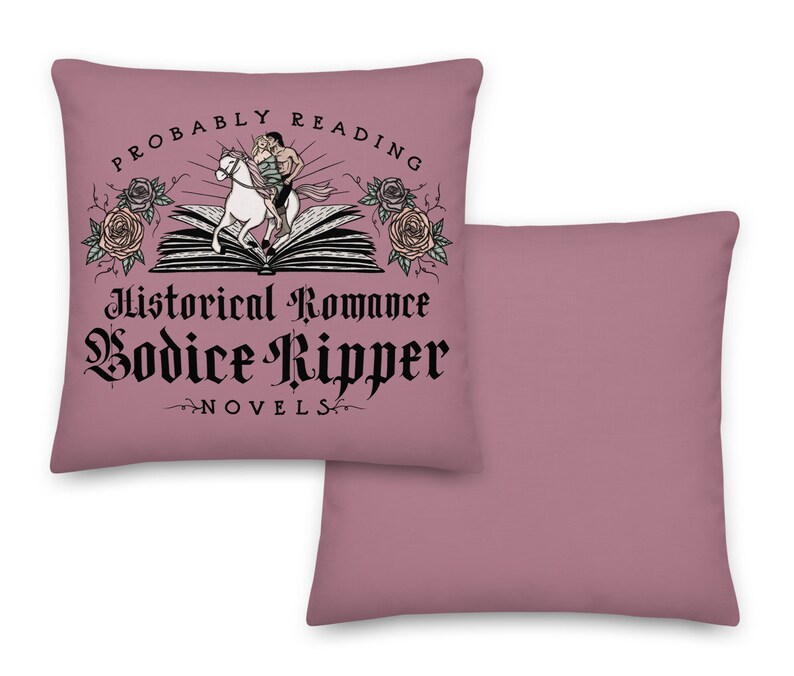 May include: A pink throw pillow featuring the text "Probably Reading Historical Romance Bodice Ripper Novels" in black lettering. The text is surrounded by a graphic of a book with a couple riding a white horse on top of it. There are roses on either side of the book.
