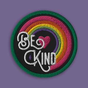 May include: A round embroidered patch with a rainbow design and the text "Be Kind" in white lettering. A small red heart is in the center of the rainbow.