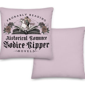 May include: A pink throw pillow with a black and white graphic design. The graphic features a couple riding a horse on top of an open book. The text reads "Probably Reading Historical Romance Bodice Ripper Novels".