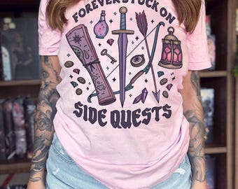 May include: A light pink t-shirt featuring a graphic print with the text "FOREVER STUCK ON SIDE QUESTS". The design includes a sword, a bow and arrow, a scroll, a potion bottle, a lantern, a key, coins, and crystals in shades of purple and pink.