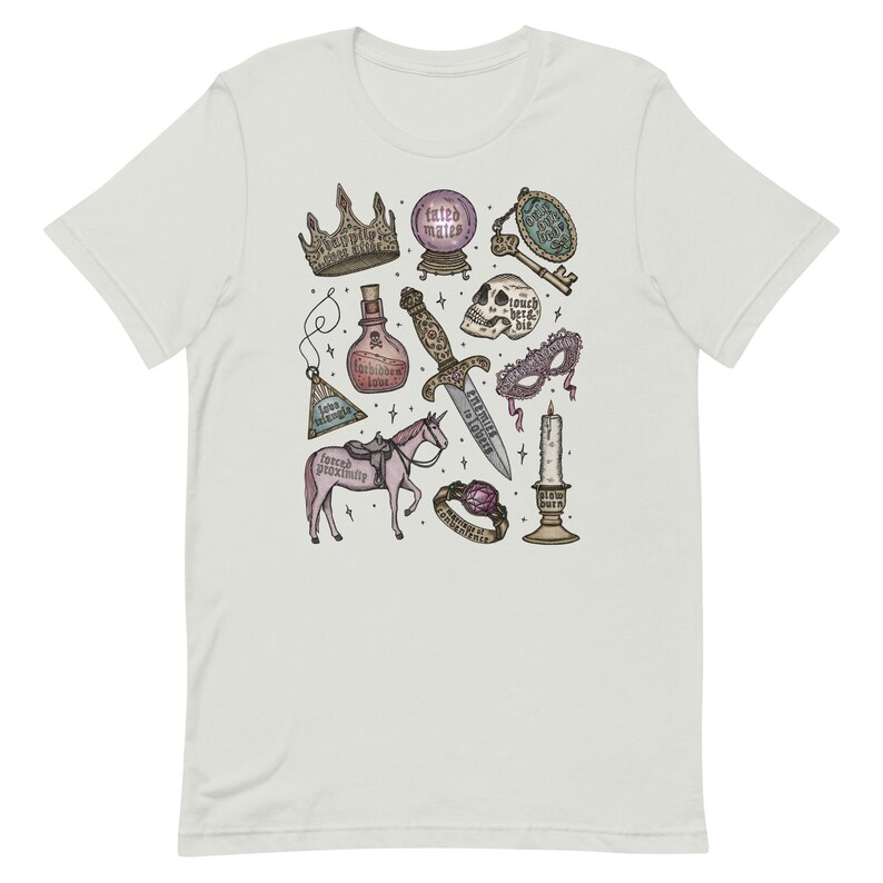 K&ouml;nnte beinhalten: Ein wei&szlig;es T-Shirt mit einer bunten Illustration eines Einhorns, einer Krone, eines Dolches, eines Sch&auml;dels, einer Zaubertrankflasche, einer Maske, eines Schl&uuml;ssels, eines Rings und einer Kerze. Der Text auf der Illustration lautet "gezwungene N&auml;he", "in Manie", "hier ber&uuml;hren, bei&szlig;en", "grausam, aber nett", "langsam brennen", "Ring der Erinnerung" und "Liebe ist ein Schlachtfeld".