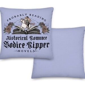 May include: A light blue throw pillow with a white design. The design features a couple riding a horse on top of an open book. The text reads "Probably Reading Historical Romance Bodice Ripper Novels".