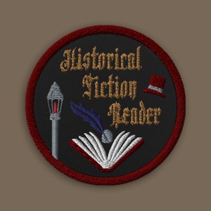 以下が含まれることがあります： 「Historical Fiction Reader」という金色の文字が書かれた黒と赤の刺繍パッチ。パッチには、羽根ペン、開いた本、シルクハットが描かれている。