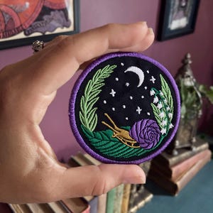 May include: A purple and green embroidered patch with a snail, a rose, and a crescent moon on a black background. The patch is surrounded by a purple border and features a green leaf design.