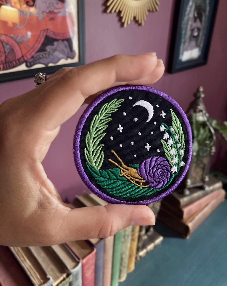 May include: A purple and green embroidered patch with a snail, a rose, and a crescent moon on a black background. The patch is surrounded by a purple border and features a green leaf design.