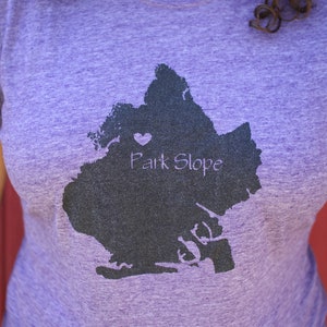 Op de afbeelding: Paars T-shirt met een zwart silhouet van de wijk Park Slope in Brooklyn, New York. Een klein hartje is op het silhouet geplaatst. De tekst "Park Slope" is onder het silhouet gedrukt.