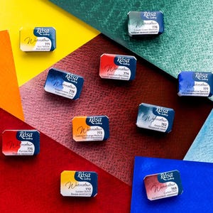 May include: A collection of watercolor paint pans from the Rosa Gallery brand, arranged on a colorful background. The pans are rectangular, with labels indicating the color names and codes. Colors include red, blue, yellow, and green.